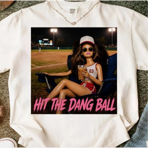 May include: White t-shirt featuring a graphic of a doll wearing a baseball cap and sunglasses, holding a drink, and sitting in a chair at a baseball field. The text "HIT THE DANG BALL" is below the image.