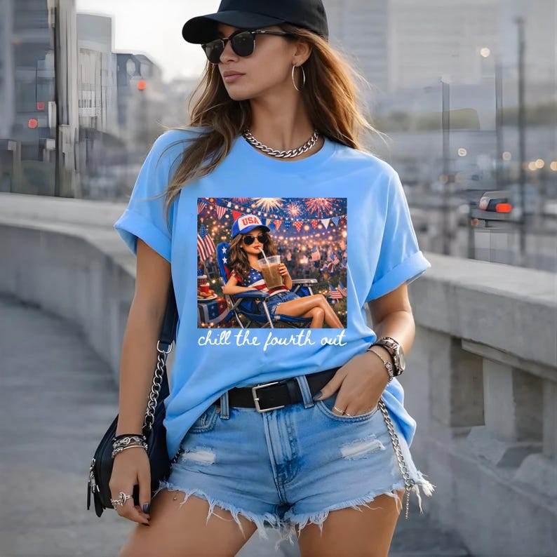 May include: Light blue t-shirt featuring a graphic of a woman relaxing with a drink, celebrating the Fourth of July. The graphic includes fireworks, flags, and the text "chill the fourth out."