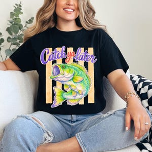 Puede incluir: Camiseta negra con una ilustraci&oacute;n colorida de un pez y el texto "Catch ya later" en morado y blanco. El pez es verde, amarillo y morado, sobre un fondo de rayas verticales naranjas.