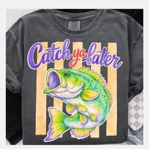 Puede incluir: Camiseta gris oscuro con una ilustraci&oacute;n colorida de un pez y el texto "Catch ya later" en morado y rojo. El dise&ntilde;o incluye rayas verticales naranjas, creando un aspecto divertido.