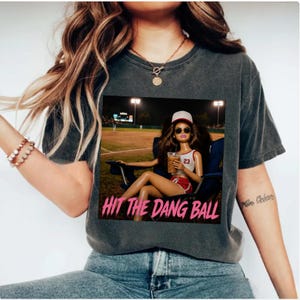 May include: Dark gray t-shirt featuring a graphic of a doll at a baseball game, with the text "HIT THE DANG BALL" in pink. The doll is wearing a baseball cap, sunglasses, and a sports uniform, holding a drink.