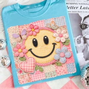May include: A light blue t-shirt featuring a patchwork design with a yellow smiley face, pink flowers, a purple butterfly, and a pink heart. The smiley face has black eyes and a stitched black smile. The patchwork includes various floral and gingham patterns.
