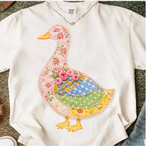 May include: White t-shirt featuring a patchwork goose design. The goose has a pink floral neck, a blue and white polka-dot body, and a yellow floral bottom. The design is outlined with pearls and has yellow feet.