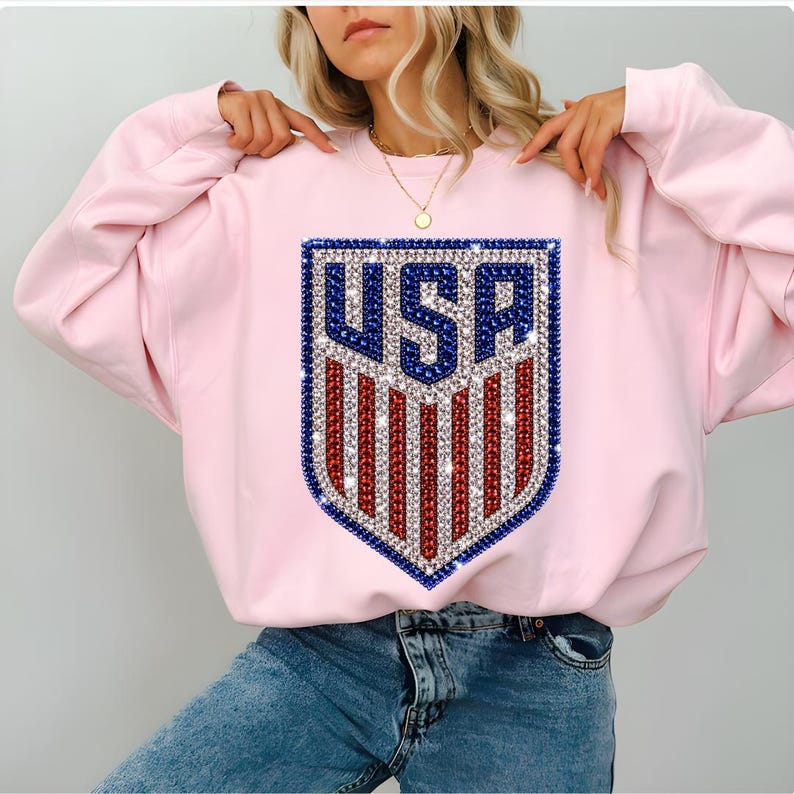 K&ouml;nnte beinhalten: Ein hellrosa Langarm-Sweatshirt mit einer zentralen schildf&ouml;rmigen Grafik und dem Text USA. Das Design ist mit funkelnden blauen, wei&szlig;en und roten Strasssteinen verziert, die in einem patriotischen Muster aus Streifen und Buchstaben angeordnet sind.