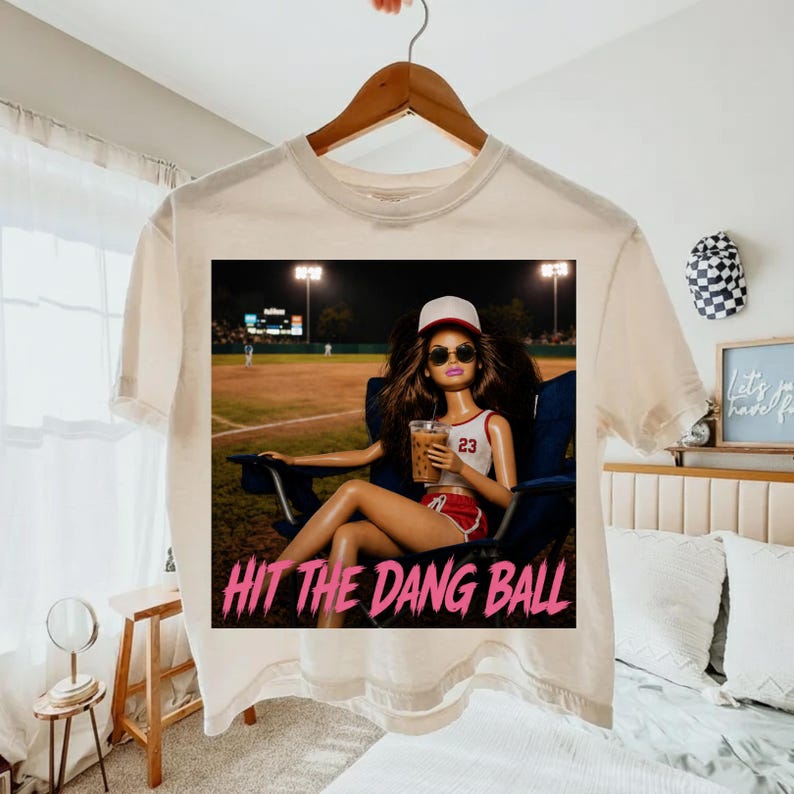 May include: Cream-colored t-shirt featuring a graphic of a doll wearing sunglasses, a baseball cap, and a sports uniform, holding a drink. The text "HIT THE DANG BALL" is printed in pink. The doll is seated in a chair at a baseball field.