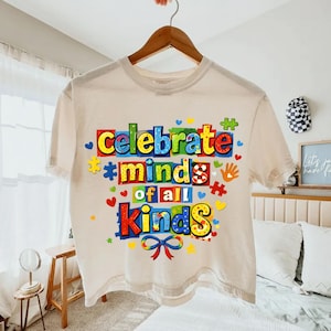 以下が含まれることがあります： 「Celebrate Minds of All Kinds」の文字がカラフルなブロック体でプリントされた、クリーム色のTシャツ。パズルのピース、ハート、手形のデザイン。木製のハンガーに掛けられています。