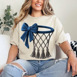 Puede incluir: Camiseta beige con un dise&ntilde;o de aro de baloncesto azul y un lazo. El aro es gris con una red y borlas. El lazo es de un tono azul m&aacute;s oscuro. La camiseta es de estilo casual.