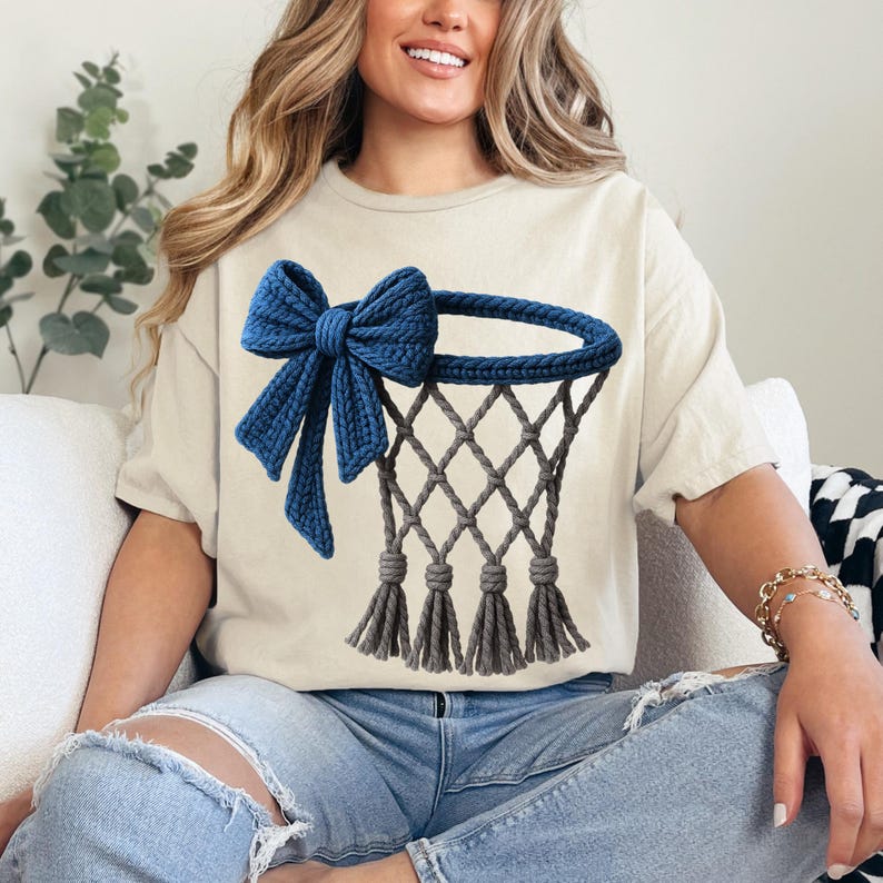 Puede incluir: Camiseta beige con un dise&ntilde;o de aro de baloncesto azul y un lazo. El aro es gris con una red y borlas. El lazo es de un tono azul m&aacute;s oscuro. La camiseta es de estilo casual.