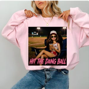May include: Light pink sweatshirt with a graphic featuring a doll in sunglasses, a baseball cap, and a drink, seated in a chair at a baseball field. The text "HIT THE DANG BALL" is below the image.