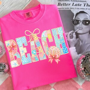 May include: A bright pink t-shirt with the word "BEACH" in a patchwork design. The letters are made of various pastel-colored fabrics, adorned with bows and seashell accents. The shirt is displayed on a pink and white checkered surface.
