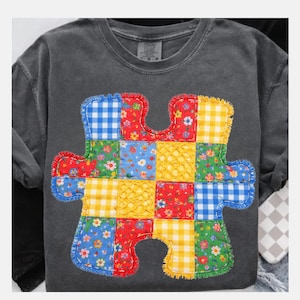 以下が含まれることがあります： 大きなカラフルなパズルピースのデザインが特徴の、濃いグレーのクルーネックTシャツ。 パズルピースは、赤、青、黄、緑のさまざまな生地の正方形でできており、花柄とギンガムチェック柄があります。 エッジは赤いフリルボーダーでトリミングされています。