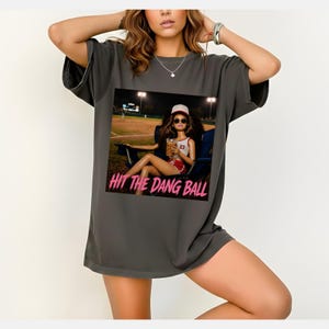 May include: Dark grey t-shirt featuring a graphic of a doll wearing a baseball cap and sunglasses, holding a drink, and sitting in a chair at a baseball field. The text "HIT THE DANG BALL" is printed in pink below the graphic.