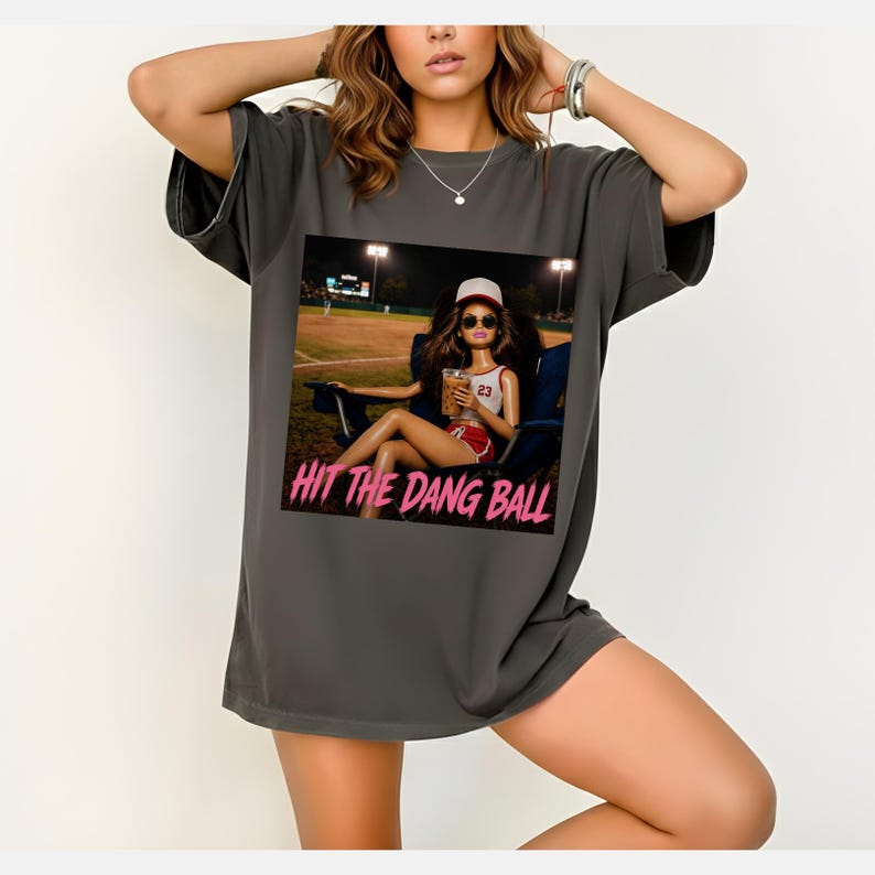 May include: Dark grey t-shirt featuring a graphic of a doll wearing a baseball cap and sunglasses, holding a drink, and sitting in a chair at a baseball field. The text "HIT THE DANG BALL" is printed in pink below the graphic.