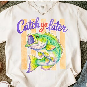 Puede incluir: Camiseta blanca con una ilustraci&oacute;n de acuarela de un pez lubina verde y amarillo. El texto "Catch ya later" est&aacute; escrito encima del pez en morado y rojo.