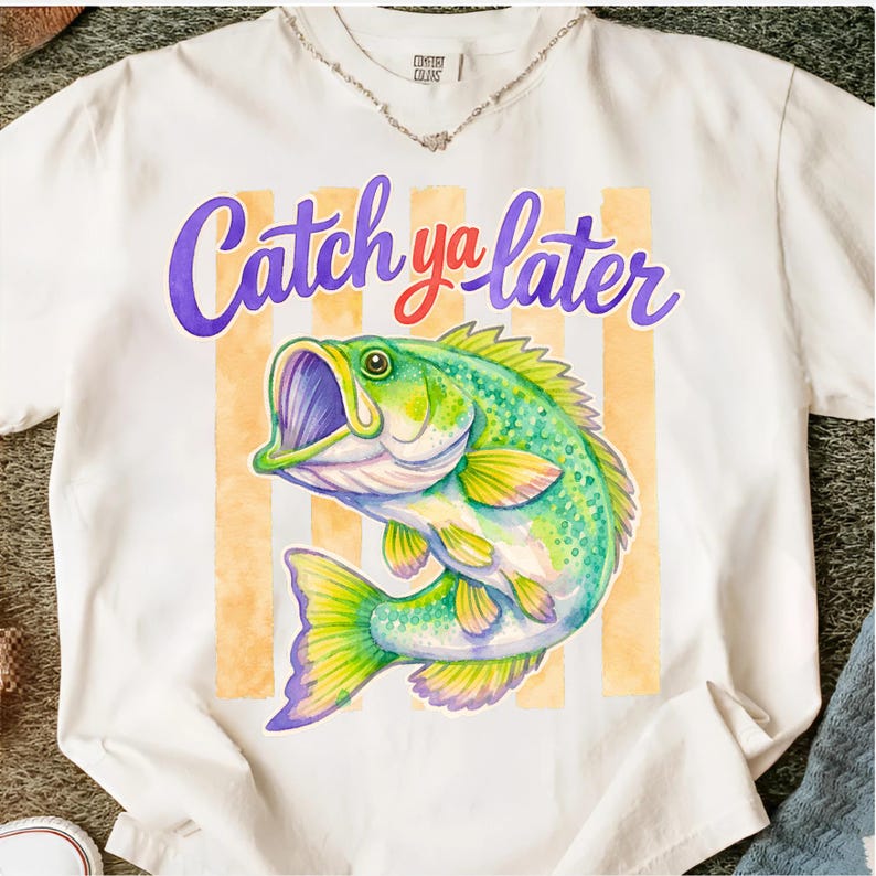 Puede incluir: Camiseta blanca con una ilustraci&oacute;n de acuarela de un pez lubina verde y amarillo. El texto "Catch ya later" est&aacute; escrito encima del pez en morado y rojo.