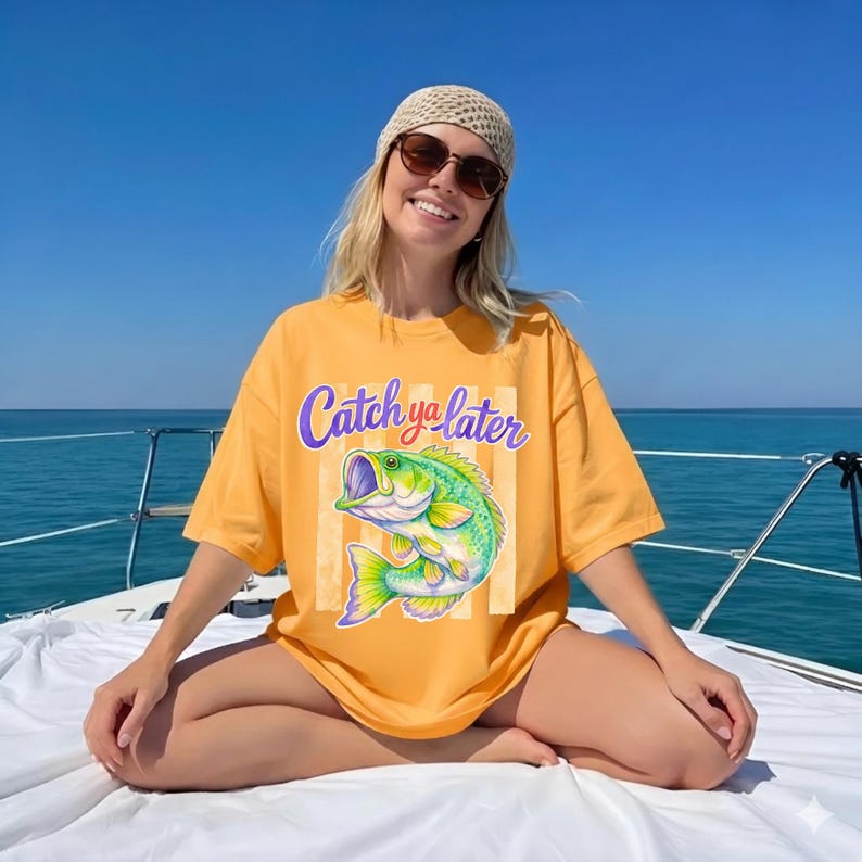 Puede incluir: Camiseta de color amarillo mostaza con el texto "Catch ya later" sobre una colorida ilustraci&oacute;n de un pez. El pez es verde, azul y amarillo. La camiseta la lleva una persona en un barco.