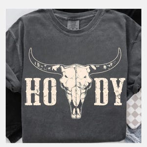 May include: Dark gray t-shirt featuring a white bull skull graphic with the word "HOODY" in a distressed font. The shirt has a crew neck and short sleeves, perfect for casual wear.