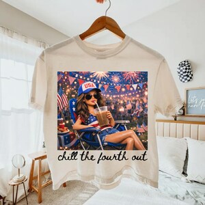 May include: Cream-colored t-shirt featuring a graphic of a woman relaxing with a drink, wearing a USA hat, and surrounded by fireworks and flags. The text "chill the fourth out" is printed below the image.