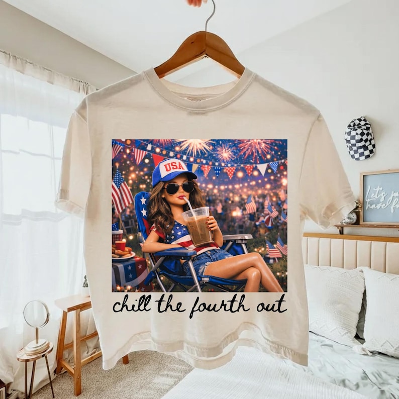 May include: Cream-colored t-shirt featuring a graphic of a woman relaxing with a drink, wearing a USA hat, and surrounded by fireworks and flags. The text "chill the fourth out" is printed below the image.