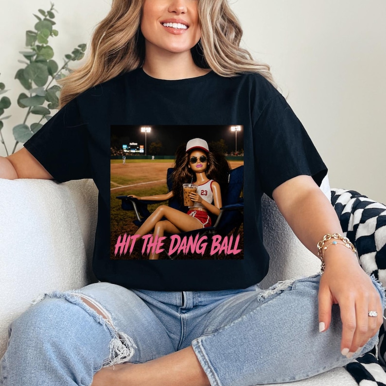 May include: Black t-shirt featuring a graphic of a doll at a baseball game, holding a drink. The doll wears a baseball cap and sunglasses. The text "HIT THE DANG BALL" is in pink. The shirt is worn by a person.
