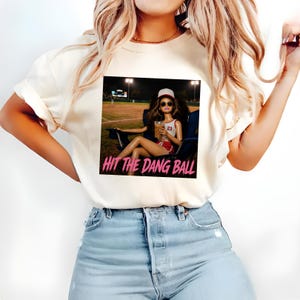 May include: Cream-colored t-shirt featuring a graphic of a doll in a baseball setting, with the text "HIT THE DANG BALL" in pink. The doll is wearing sunglasses, a baseball cap, and a sports uniform. The shirt is paired with blue jeans.