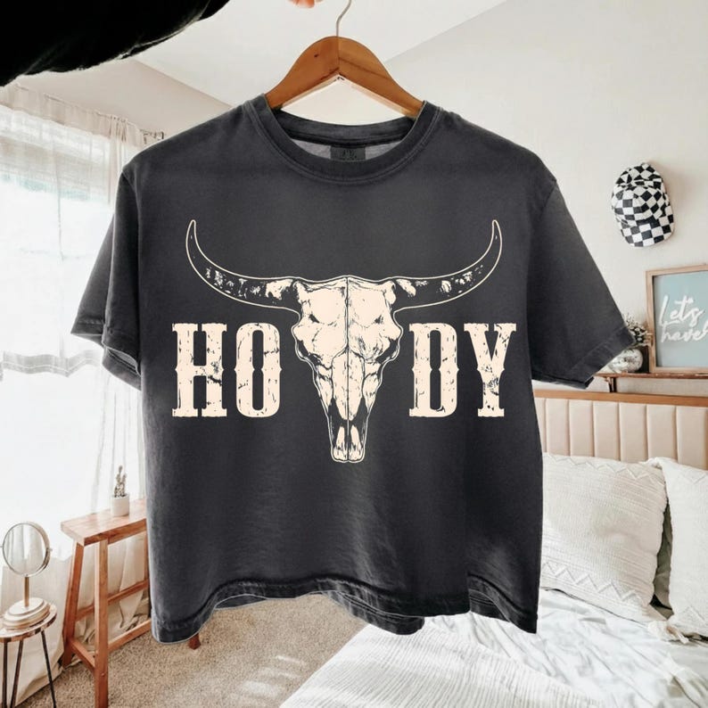 May include: Dark gray t-shirt featuring a white bull skull graphic with the word "HOWDY" in a distressed font. The shirt is hanging on a wooden hanger, showcasing a Western or country theme.