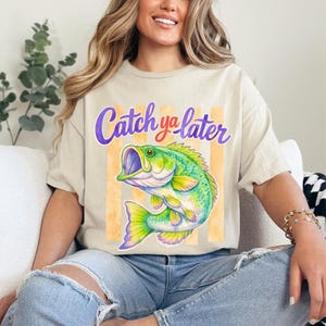 Puede incluir: Camiseta beige con una ilustraci&oacute;n colorida de un pez y el texto "Catch ya later" en morado y rojo. El pez es verde, amarillo y azul, con la boca abierta. El fondo tiene rayas verticales naranjas.
