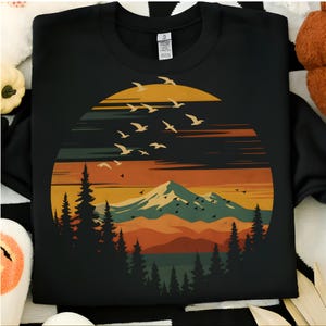 May include: Black sweatshirt with a nature-themed design. The graphic depicts a sunset, mountains, pine trees, and birds in flight. The colour scheme includes orange, yellow, green, and brown hues.