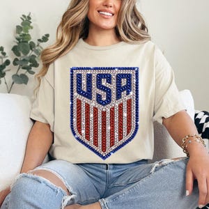 K&ouml;nnte beinhalten: Ein cremefarbenes Kurzarm-T-Shirt mit einer gro&szlig;en schildf&ouml;rmigen Grafik auf der Brust. Die Grafik zeigt den Text USA in blauen Strasssteinen &uuml;ber vertikalen roten und wei&szlig;en Strassstreifen, alles umrandet von einem Rahmen aus blauen Strasssteinen.