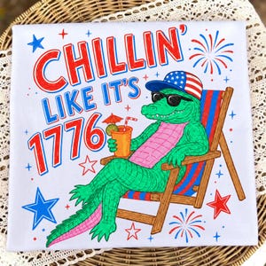 Può includere: Maglietta bianca con un alligatore dei cartoni animati con occhiali da sole e un cappello patriottico, che si rilassa su una sedia a sdraio. La maglietta presenta la scritta "CHILLIN' LIKE IT'S 1776" in rosso e blu, con fuochi d'artificio e stelle.