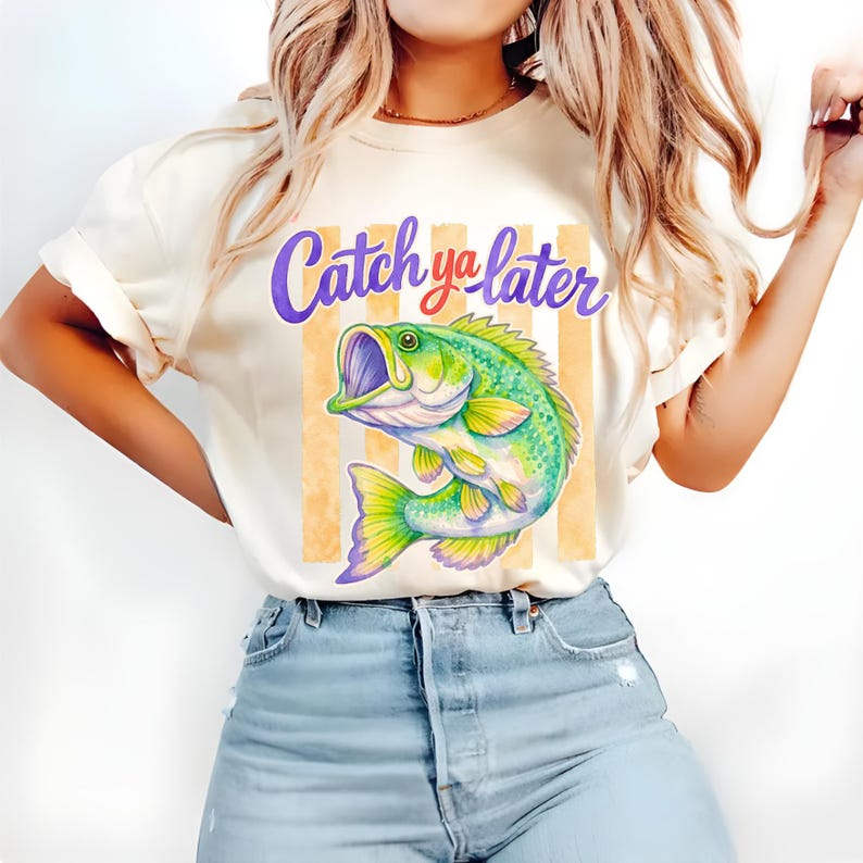 Puede incluir: Camiseta color crema con una colorida ilustraci&oacute;n de un pez verde y amarillo con la boca abierta. El texto "Catch ya later" est&aacute; escrito encima del pez en escritura morada y roja. Hay rayas verticales naranjas detr&aacute;s del pez.