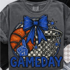 Puede incluir: Camiseta gris oscuro con un diseño de baloncesto. El diseño incluye una pelota de baloncesto naranja, un lazo azul, un dedo de espuma, estrellas y la palabra "GAMEDAY" en azul. La camiseta tiene cuello redondo y mangas cortas.
