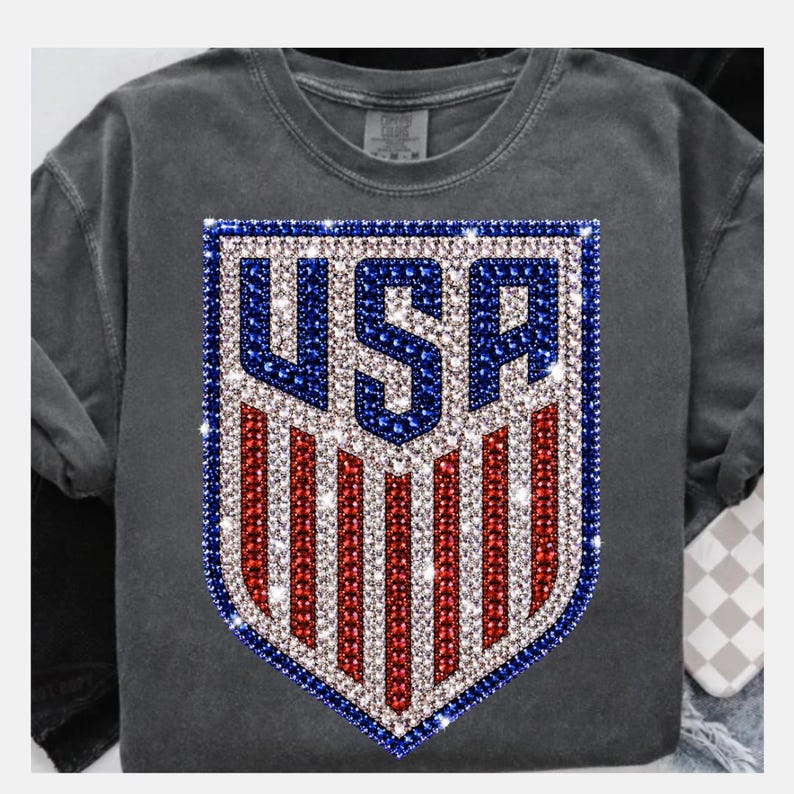 K&ouml;nnte beinhalten: Ein anthrazitfarbenes T-Shirt mit einem zentralen schildf&ouml;rmigen Motiv aus funkelnden roten, wei&szlig;en und blauen Strasssteinen. Das Design zeigt die Buchstaben USA in Blau vor einem Hintergrund aus vertikalen roten und wei&szlig;en Streifen.