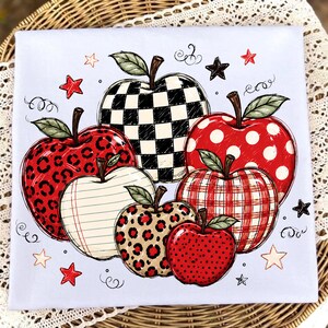 May include: A light blue fabric square featuring a graphic illustration of six apples with various patterns including black and white checkerboard, red leopard print, white lined paper, red polka dots, red plaid, and red with black dots, accented by small stars.