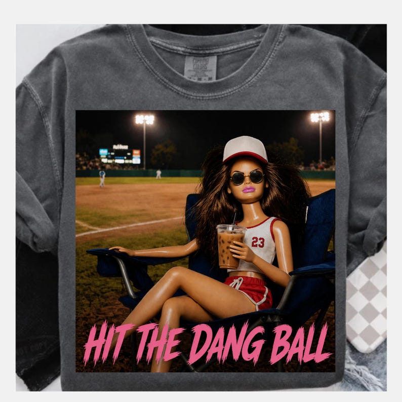 May include: Dark gray t-shirt featuring a graphic of a doll wearing sunglasses and a baseball cap, holding a drink, and sitting in a chair at a baseball field. The text "HIT THE DANG BALL" is printed in pink below the graphic.