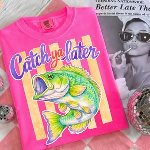 Puede incluir: Una camiseta rosa brillante con la frase "Catch ya later" en una escritura p&uacute;rpura. Una colorida ilustraci&oacute;n de un pez verde y amarillo est&aacute; debajo del texto. La camiseta tiene cuello redondo y mangas cortas. La camiseta est&aacute; sobre una superficie a cuadros rosa y blanco.