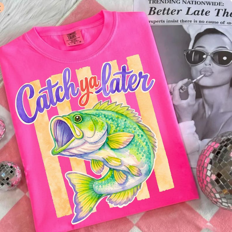 Puede incluir: Una camiseta rosa brillante con la frase "Catch ya later" en una escritura p&uacute;rpura. Una colorida ilustraci&oacute;n de un pez verde y amarillo est&aacute; debajo del texto. La camiseta tiene cuello redondo y mangas cortas. La camiseta est&aacute; sobre una superficie a cuadros rosa y blanco.