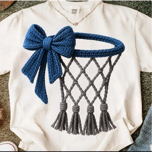 Puede incluir: Camiseta blanca con un lazo azul tejido y un dise&ntilde;o de red de baloncesto gris tejida. El lazo es grande y detallado, con extremos largos y fluidos. La red tiene una apariencia texturizada con borlas en la parte inferior.