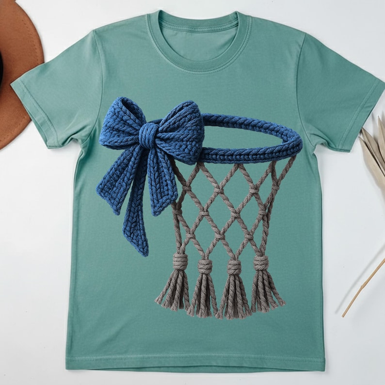 Puede incluir: Camiseta verde azulado con un dise&ntilde;o de ganchillo de una canasta de baloncesto. La canasta tiene un lazo azul, una red gris y borlas grises. El lazo y el aro son de color azul oscuro. El dise&ntilde;o es detallado.