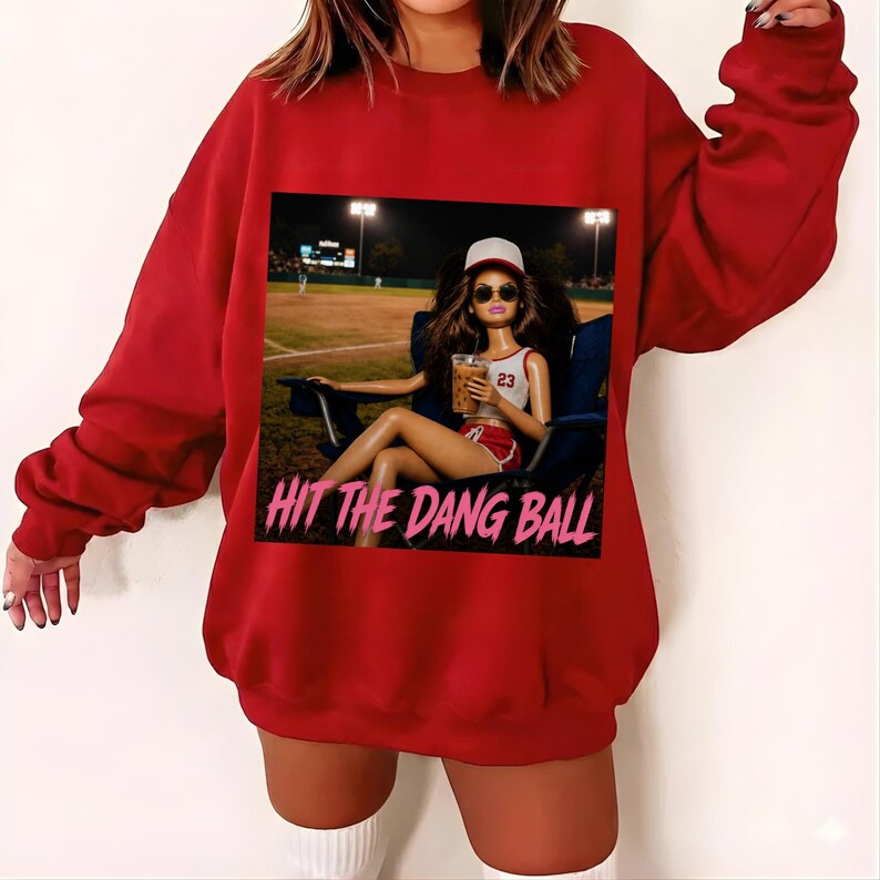 May include: A red sweatshirt featuring a graphic of a doll wearing sunglasses and a baseball cap, holding a drink, and sitting in a chair at a baseball field. The text "HIT THE DANG BALL" is below the graphic.