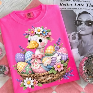 Puede incluir: Una camiseta rosa brillante con una ilustración de un ganso en una cesta con huevos de Pascua y flores. El ganso lleva una corona de flores y un lazo rosa. La camiseta tiene la etiqueta de la marca Comfort Colors.