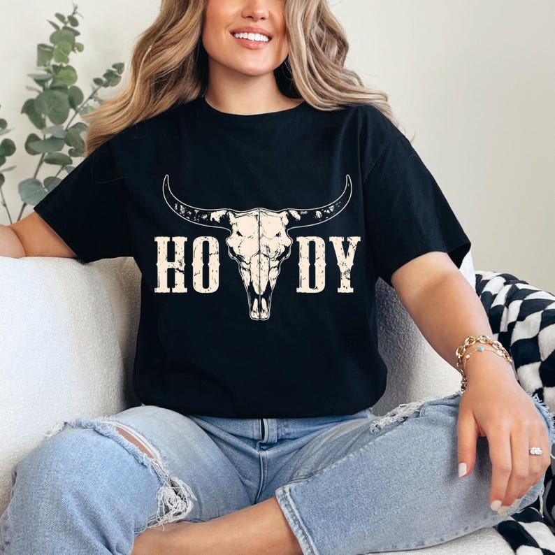May include: Black t-shirt featuring a cream-colored bull skull graphic with the word "HOWDY" in a bold, western-style font. The shirt is a relaxed fit, perfect for casual wear. The design has a vintage, rustic aesthetic.