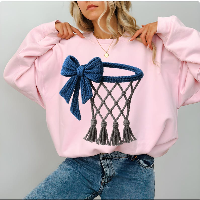 Puede incluir: Una sudadera rosa claro con un lazo azul tejido y un dise&ntilde;o de red de baloncesto gris. La red tiene un patr&oacute;n de diamantes y borlas grises. La sudadera es de estilo casual y c&oacute;modo.