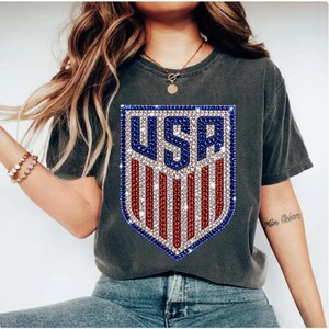 K&ouml;nnte beinhalten: Ein anthrazitfarbenes Kurzarm-T-Shirt mit einer gro&szlig;en schildf&ouml;rmigen Grafik auf der Brust. Die Grafik zeigt den Text USA in Blau, darunter rote und wei&szlig;e vertikale Streifen, alles in einer funkelnden, strassartigen Textur dargestellt.
