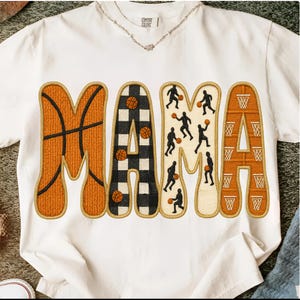 May include: White t-shirt featuring the word "MAMA" in a basketball design. The letters are decorated with basketballs, hoops, and player silhouettes. The shirt has a crew neck and a comfortable fit, suitable for casual wear.