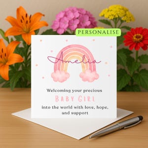 Puede incluir: Una tarjeta de felicitación con un diseño de arcoíris en acuarela, el nombre "Amelia" en escritura cursiva y el texto "Welcoming your precious BABY GIRL." La tarjeta está sobre un sobre blanco, con un bolígrafo. Flores en naranja, rosa, amarillo y rojo en el fondo.