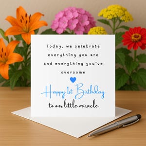 Puede incluir: Una tarjeta de felicitación blanca con el texto azul "Happy 1st Birthday to our little miracle". La tarjeta está sobre un sobre blanco, con un bolígrafo plateado. El fondo incluye lirios naranjas, flores rosas y amarillas y una zinnia roja.