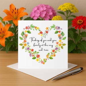 Könnte beinhalten: Eine weiße Beileidskarte mit einem herzförmigen Blumenkranz. Auf der Karte steht: „In Gedanken bei Ihnen und Ihrer Familie in dieser sehr traurigen Zeit“. Die Karte liegt auf einem weißen Umschlag, mit einem silbernen Stift auf dem Tisch.