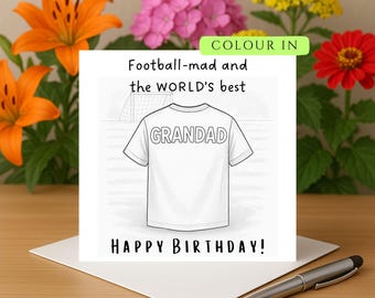 Op de afbeelding: Een witte verjaardagskaart met de tekst "Football-mad and the WORLD'S best GRANDAD HAPPY BIRTHDAY!" toont een lijntekening van een t-shirt. De kaart ligt op een houten oppervlak, met een pen en bloemen op de achtergrond.