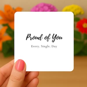 May include: A white square card with rounded corners, held by a hand with pink nail polish. The card features the text "Proud of You" in a cursive font, with "Every. Single. Day" below it.